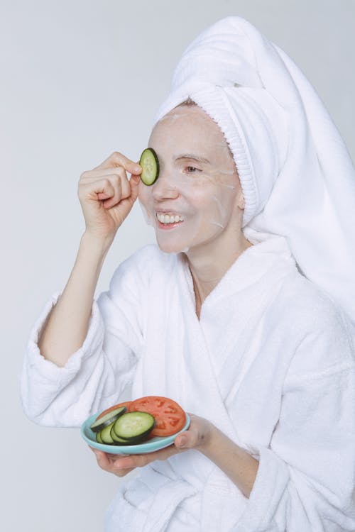 Positive young female in white towel and bathrobe enjoying healthy skin care morning routine with cucumber slices and sheet mask