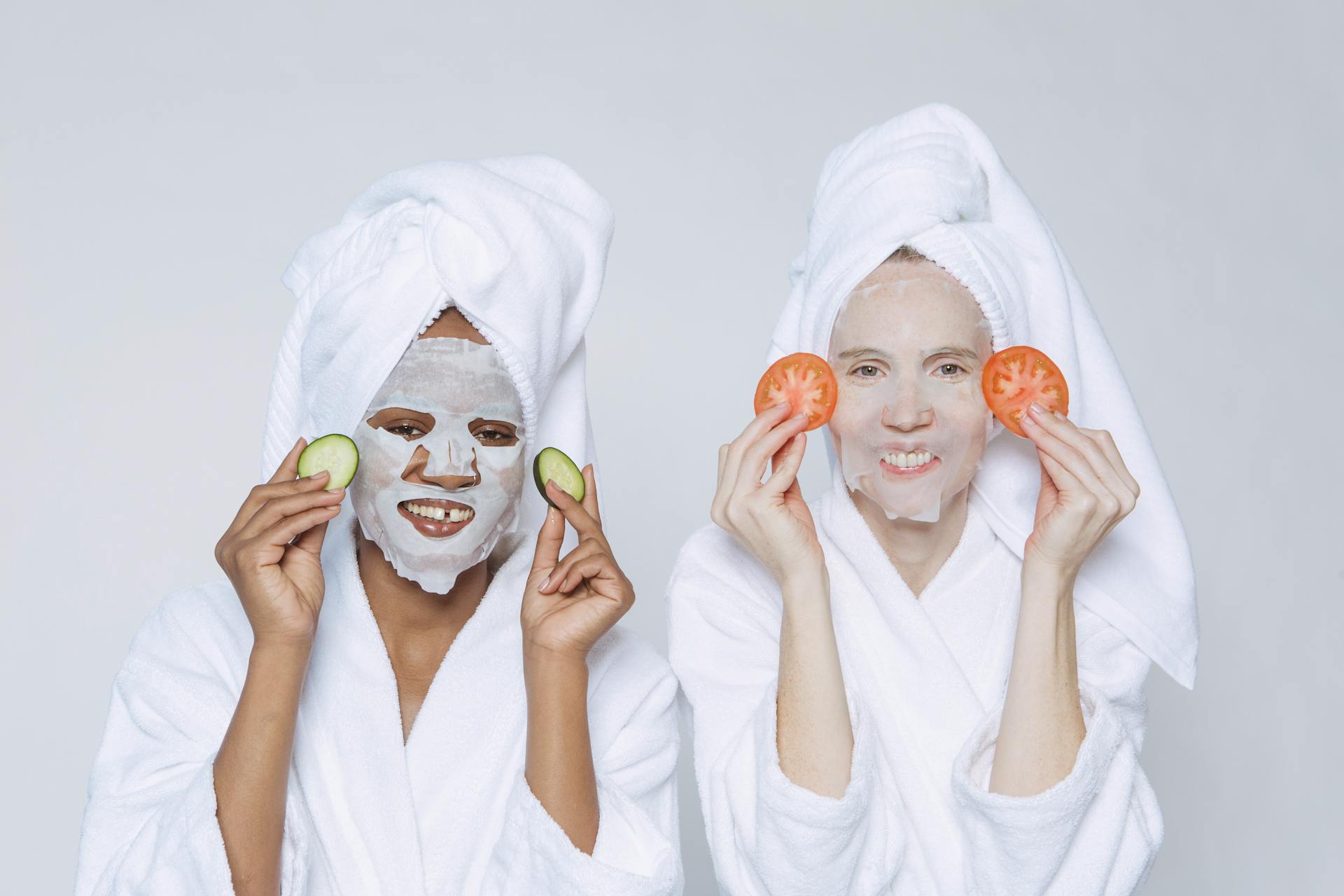 Optimistic multiethnic females wearing towels and bathrobes applying slices of cucumbers and tomatoes for skincare moisturizing treatment