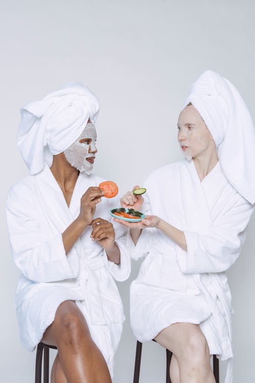 Free Multiethnic females in bathrobes and towels moisturizing faces with wet sheet masks and taking slices of tomato and cucumber from plate Stock Photo