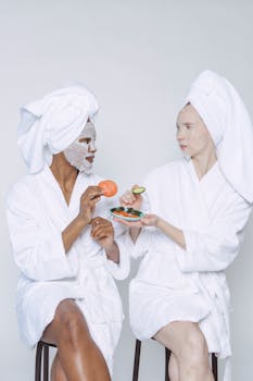 Multiethnic females in bathrobes and towels moisturizing faces with wet sheet masks and taking slices of tomato and cucumber from plate