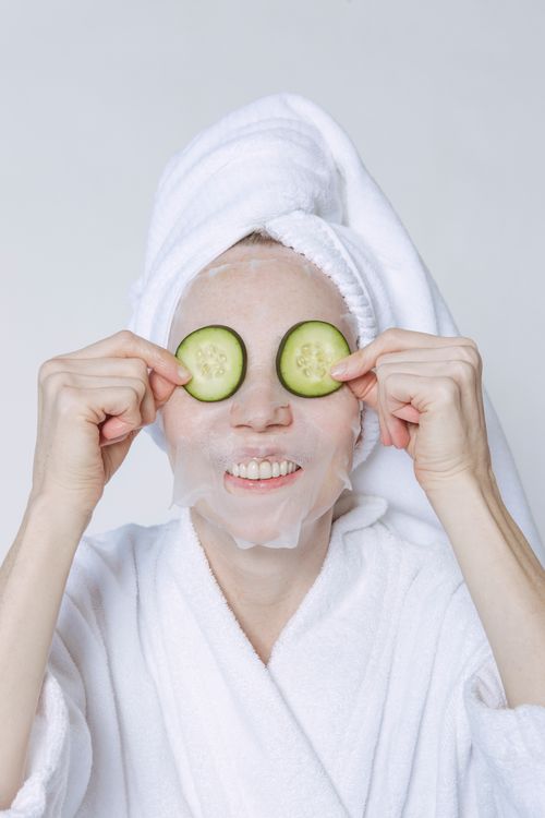 Free Positive female in towel turban and bathrobe applying cucumber slices on face with sheet mask for skincare treatment Stock Photo