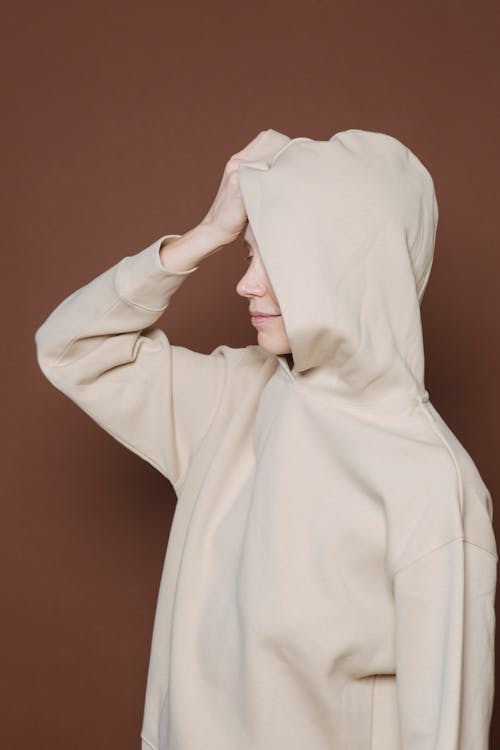 Young female model covering face with hood of oversize sweatshirt while standing against brown background