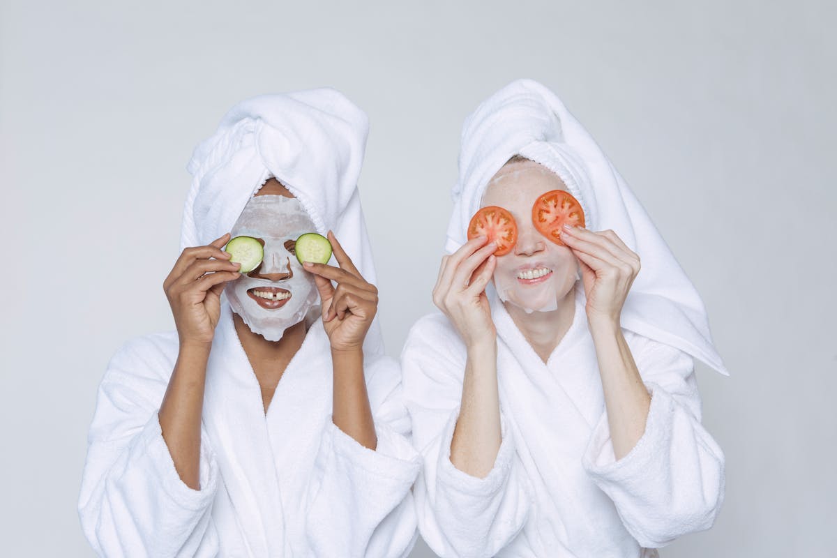 Positive diverse women in bathrobes and turbans moisturizing faces with sheet mask and applying slices of cucumber and tomato on eyes against white background