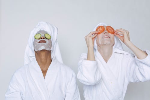 Young multiethnic females wearing bathrobes and towels moisturizing face with masks and slices of tomatoes and cucumbers against white background