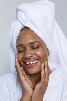 Positive African American female with closed eyes in white bathrobe and towel on head gently touching face while doing skin care routine