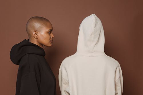 Unemotional bald African American female in oversize hoodie standing near person in similar outfit against brown background