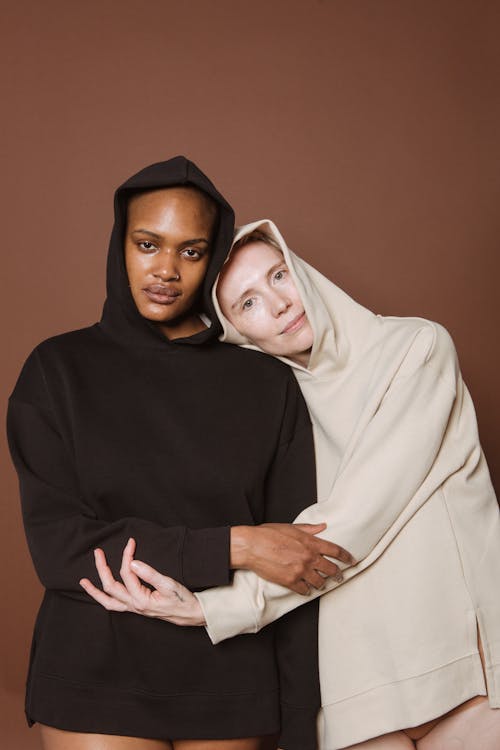 Delicate female leaning on shoulder of African American woman in hoodie while looking at camera against brown background