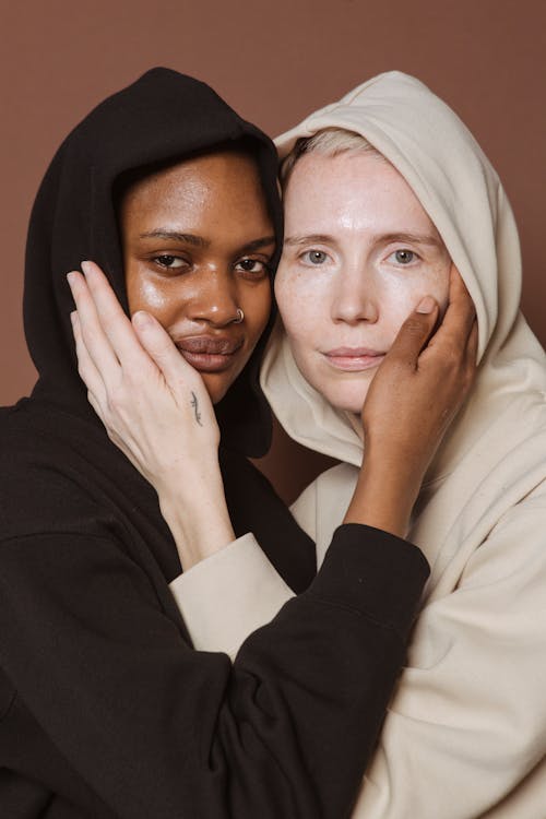 Free Multiracial female models in casual hoodies looking at camera while touching faces of each other on brown background in studio Stock Photo