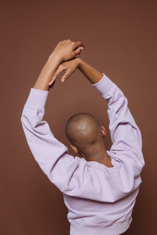 Back view of anonymous African American person in stylish wear standing on brown background with raised arms in light studio