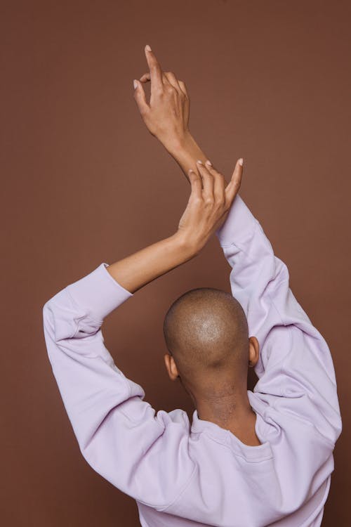 Back view of faceless African American model in casual wear standing with raised arms on brown background in light modern studio