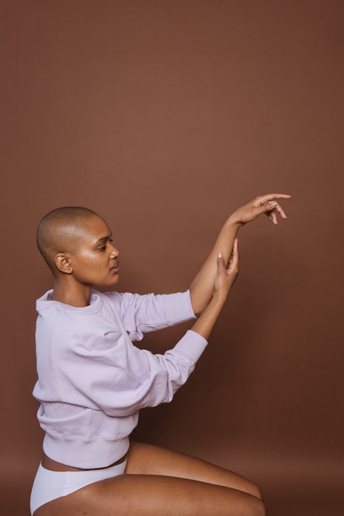 Free Side view of graceful bald young African American female model in panties and sweatshirt sitting on knees against brown background Stock Photo