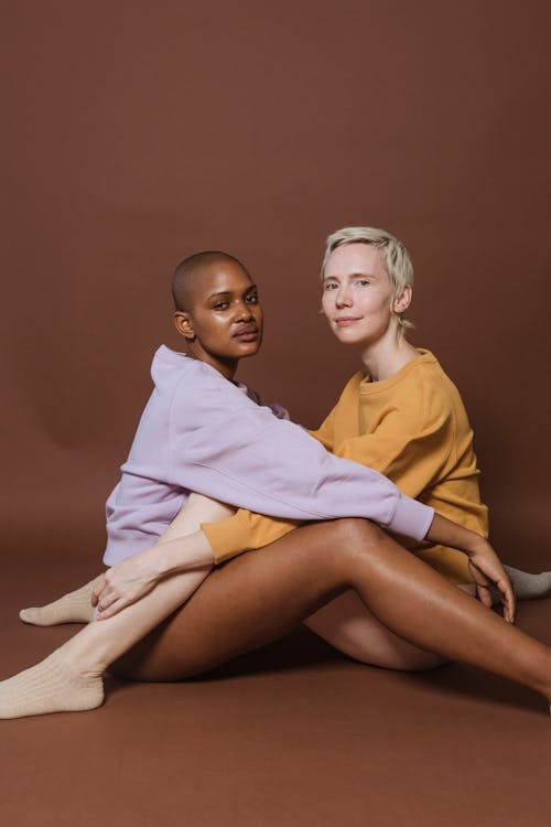 Side view of young multiracial female models in stylish underwear and sweatshirts sitting in front of each other and looking at camera against brown background