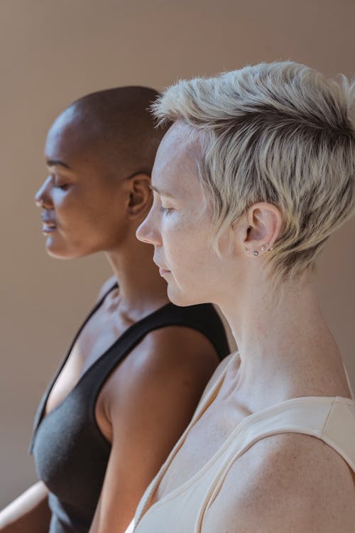 Side view of gracious young multiethnic female models in white and black bodysuits standing in brown studio and looking away pensively