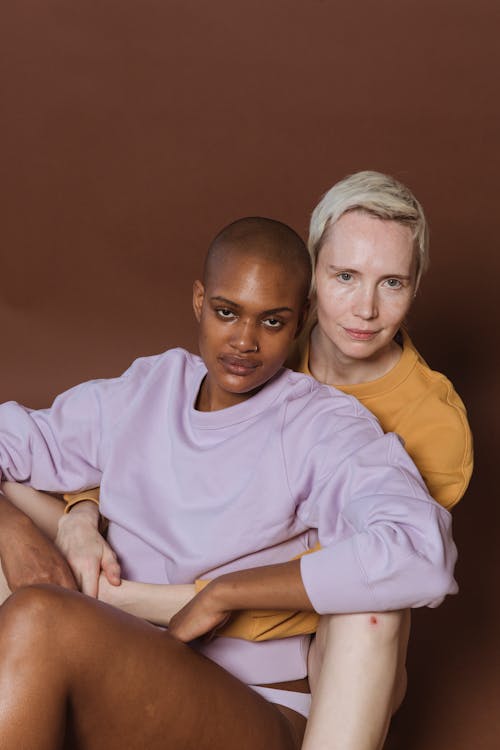 Confident adult multiracial female friends in casual clothes sitting and embracing while looking at camera in light room on brown background