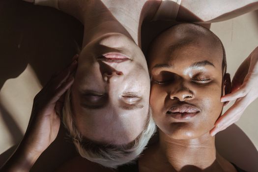 Top view of concentrated adult multiethnic women with closed eyes caressing cheeks of each other while lying on floor