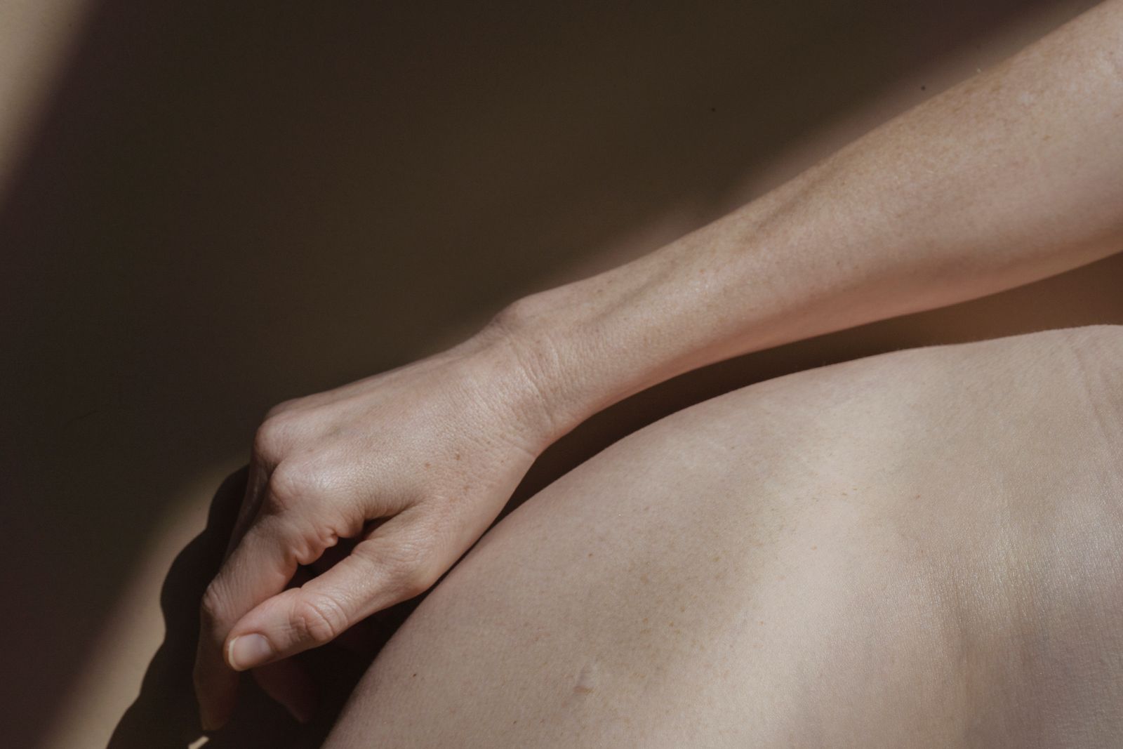 Close-up of a woman's bare forearm and hand resting on a bare shoulder in warm sunlight and shadow