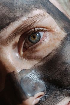 Crop woman with blue eyes and rejuvenating clay mask on face looking at camera in sunlight