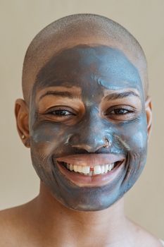 Cheerful African American female with bald head and natural clay mask on face looking at camera against beige background in studio