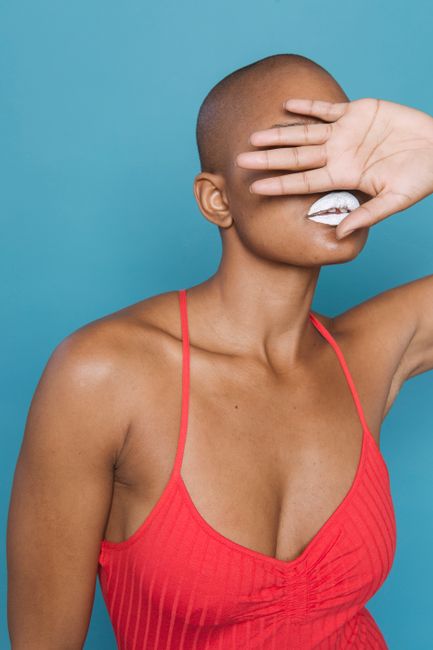 Calm bald African American female in red underwear with white lips covering eyes with hand on blue background in studio