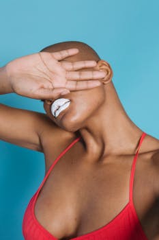 A bald woman in a red top covers her face with hand, showcasing bold makeup against a blue backdrop.