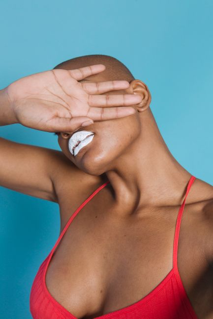 Sensual bald female in pink underwear with white lips covering face with hand against light blue background
