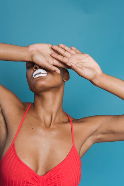 Crop African American hairless female in pink swimsuit with white lips covering eyes with hands against blue background