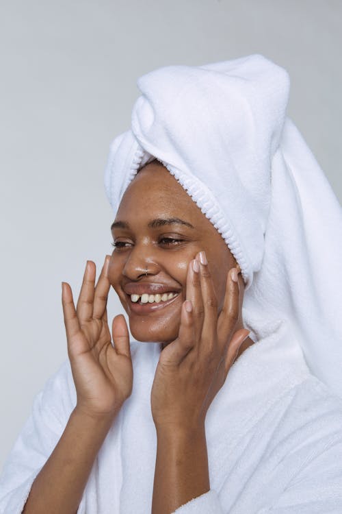 Free Side view of smiling black female in white bathrobe and towel on head smearing cream on face and looking away against white background Stock Photo