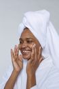 Side view of smiling black female in white bathrobe and towel on head smearing cream on face and looking away against white background