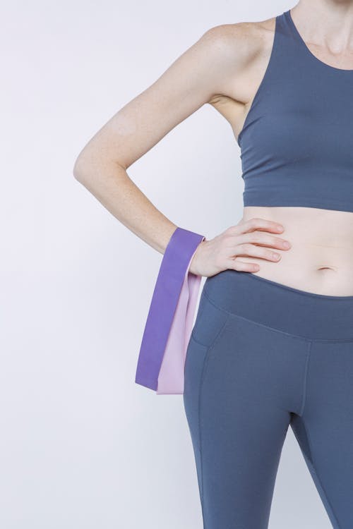 Crop unrecognizable slim female wearing purple sportswear standing with hand on waist holding resistance bands against white background