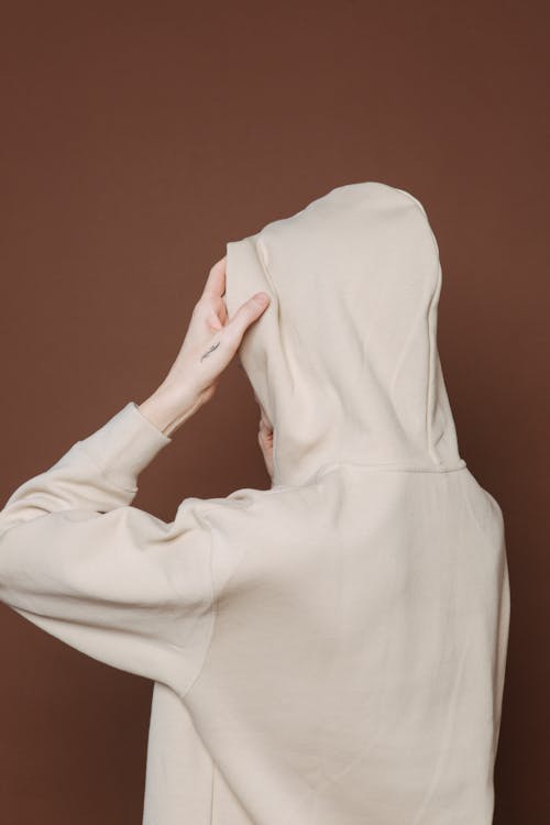 Back view of anonymous female in white hoodie standing holding hood with hands near brown background