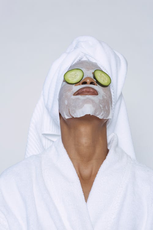 Free Unrecognizable black female in white bathrobe and towel on head and with sheet mask and cucumber slices on eyes standing head thrown back against white background Stock Photo