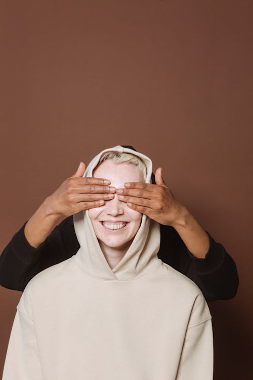 Ethnic person covering with hands eyes of smiling blond model in hoody against brown background