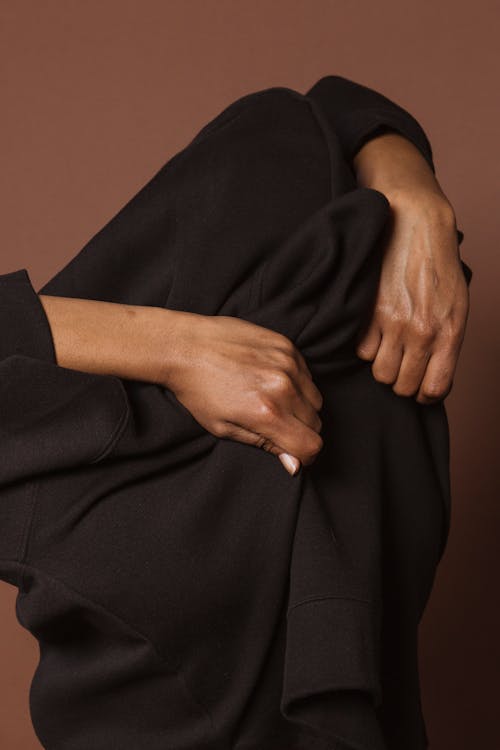 Unrecognizable black person putting on dark shirt with long sleeves against brown background in studio
