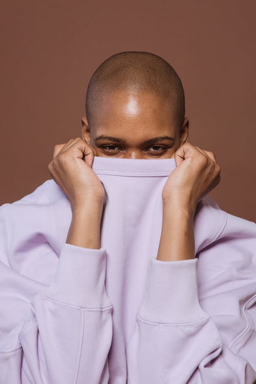 Free Black hairless woman looking at camera while covering face with pink long sleeve shirt in studio Stock Photo