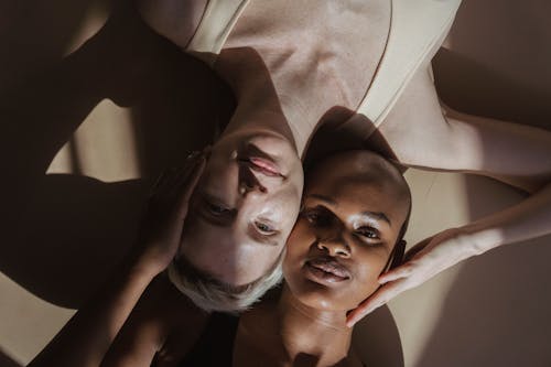 Top view of smiling multiethnic female models without makeup lying on gray surface and touching heads holding hands on face while looking at camera with shadows on face