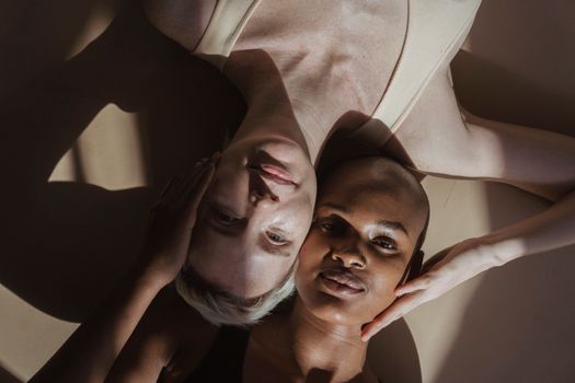 Top view of smiling multiethnic female models without makeup lying on gray surface and touching heads holding hands on face while looking at camera with shadows on face