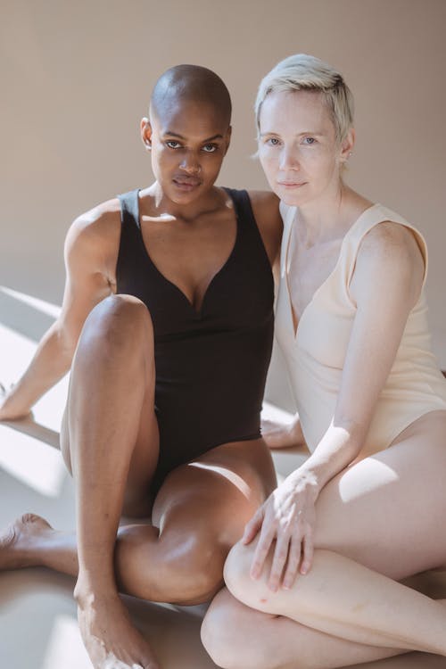 Calm diverse female models in white and black bodysuits sitting on floor and leaning on hands while looking at camera in light studio