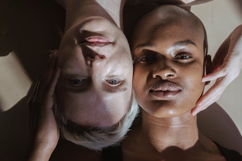 Top view of emotionless multiracial female models lying on floor touching face and looking at camera in daylight