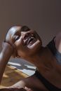 Portrait of African American hairless smiling female with closed eyes lying on floor and leaning on elbow against gray wall