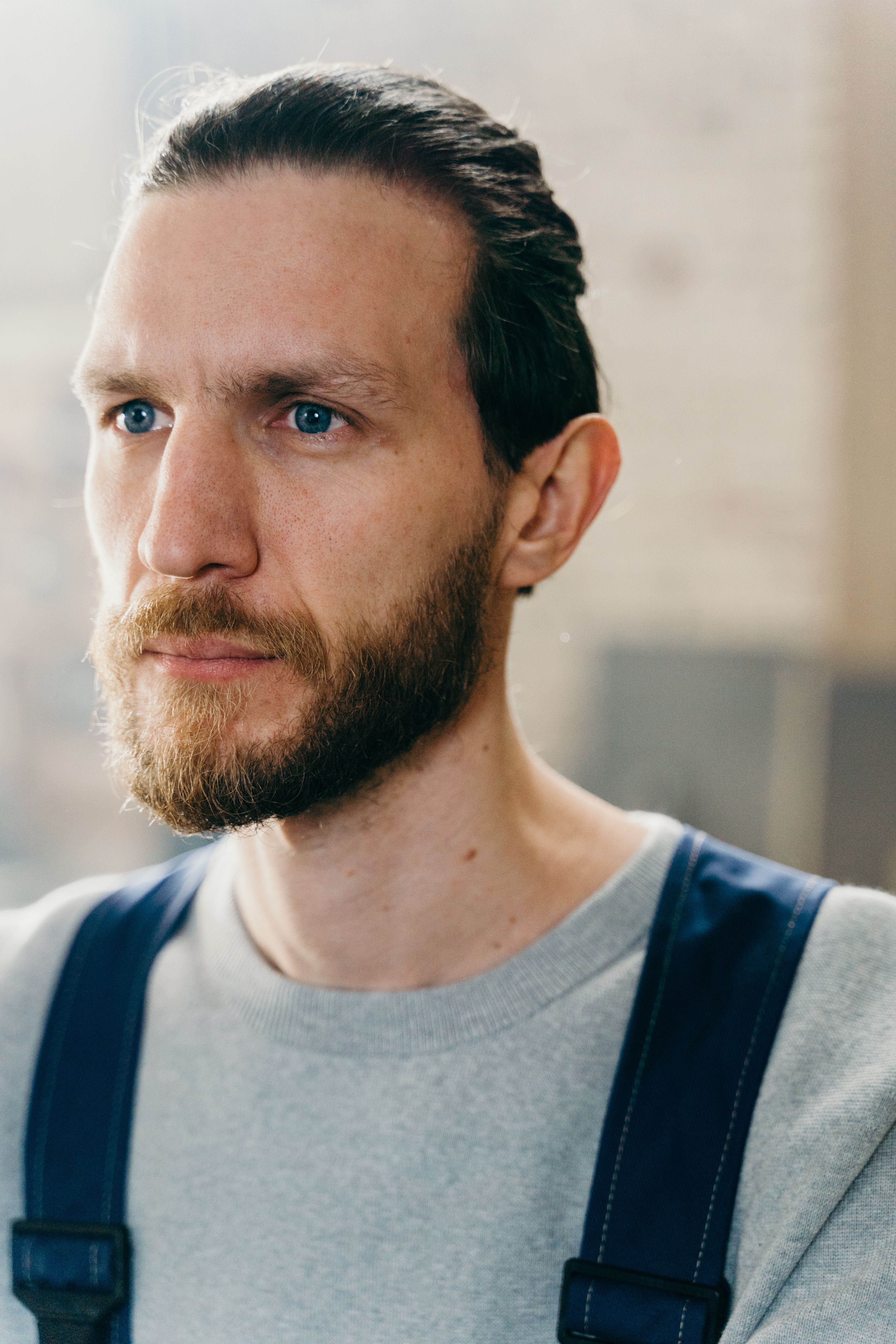 Portrait of a bearded craftsman with blue eyes and dark hair indoors wearing a gray shirt and denim apron.