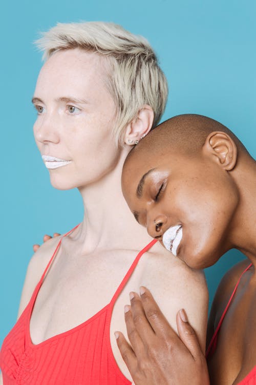 Free Bald black lady with white lipstick closing eyes while demonstrating tender feeling with blond female partner on blue background Stock Photo