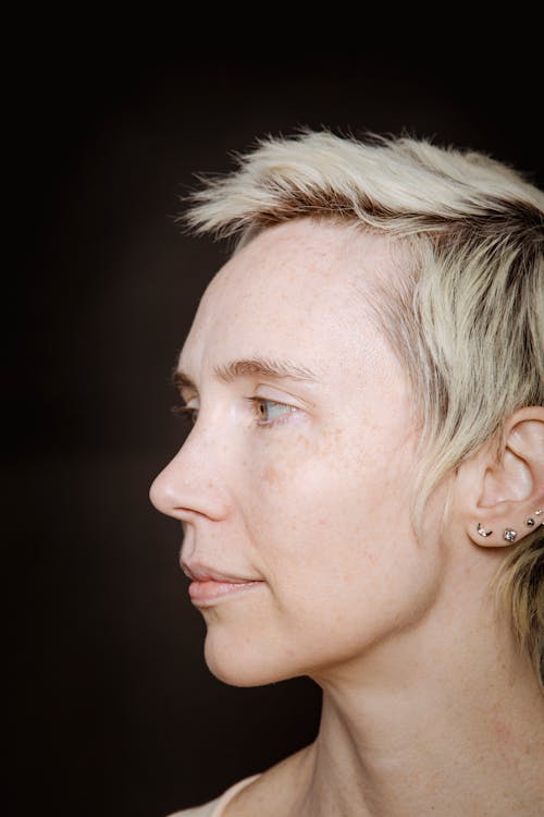 Free Side view of short haired blond female with freckles on face and ear piercing looking away against black background Stock Photo