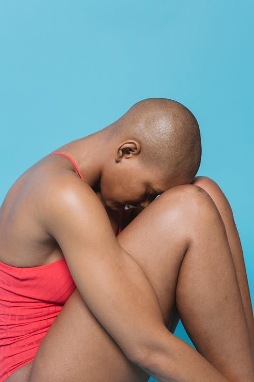 Free Side view of hairless black lady in red underwear burying face in bare knees while sitting on blue background Stock Photo