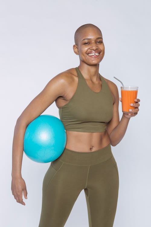 Cheerful African American lady in activewear with blue ball drinking orange juice and looking at camera against white background
