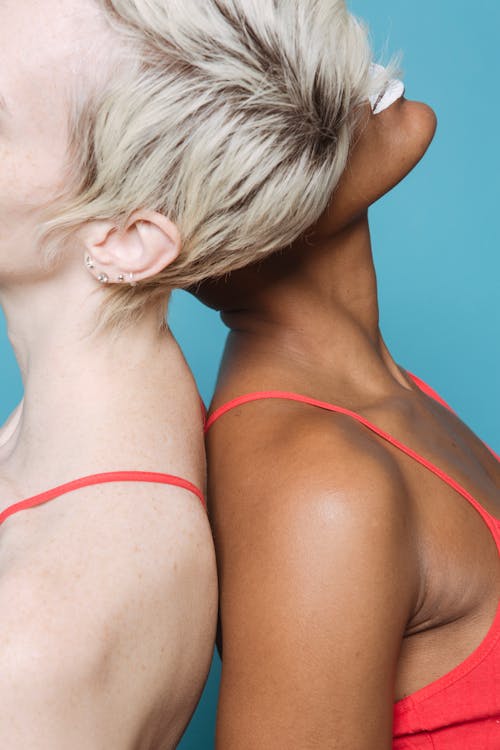 Free Side view of crop unrecognizable multiracial female models in similar light tops standing back to back against blue background in studio Stock Photo