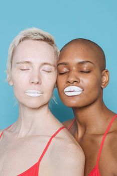 Calm young diverse female models with white lipsticks in similar tops standing together against blue background with closed eyes