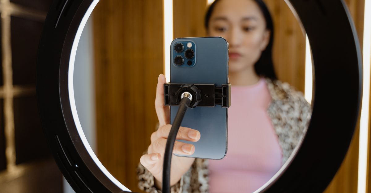 Young woman using a smartphone held by a ring light for video recording indoors.