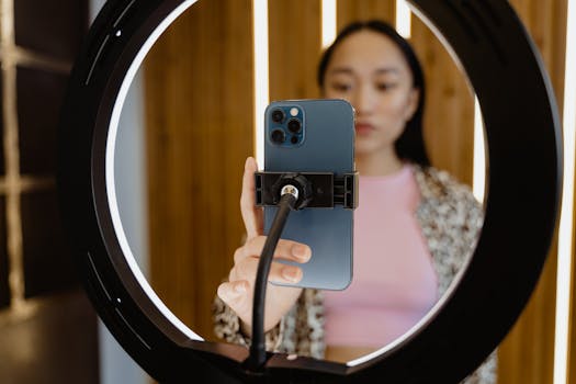 Young woman using a smartphone held by a ring light for video recording indoors.