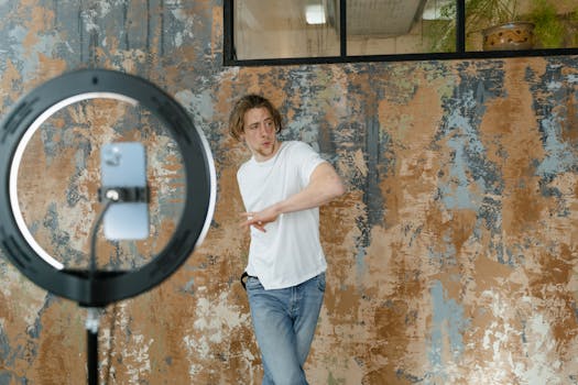Young man in casual attire dances while recording a video indoors with a smartphone and ring light.