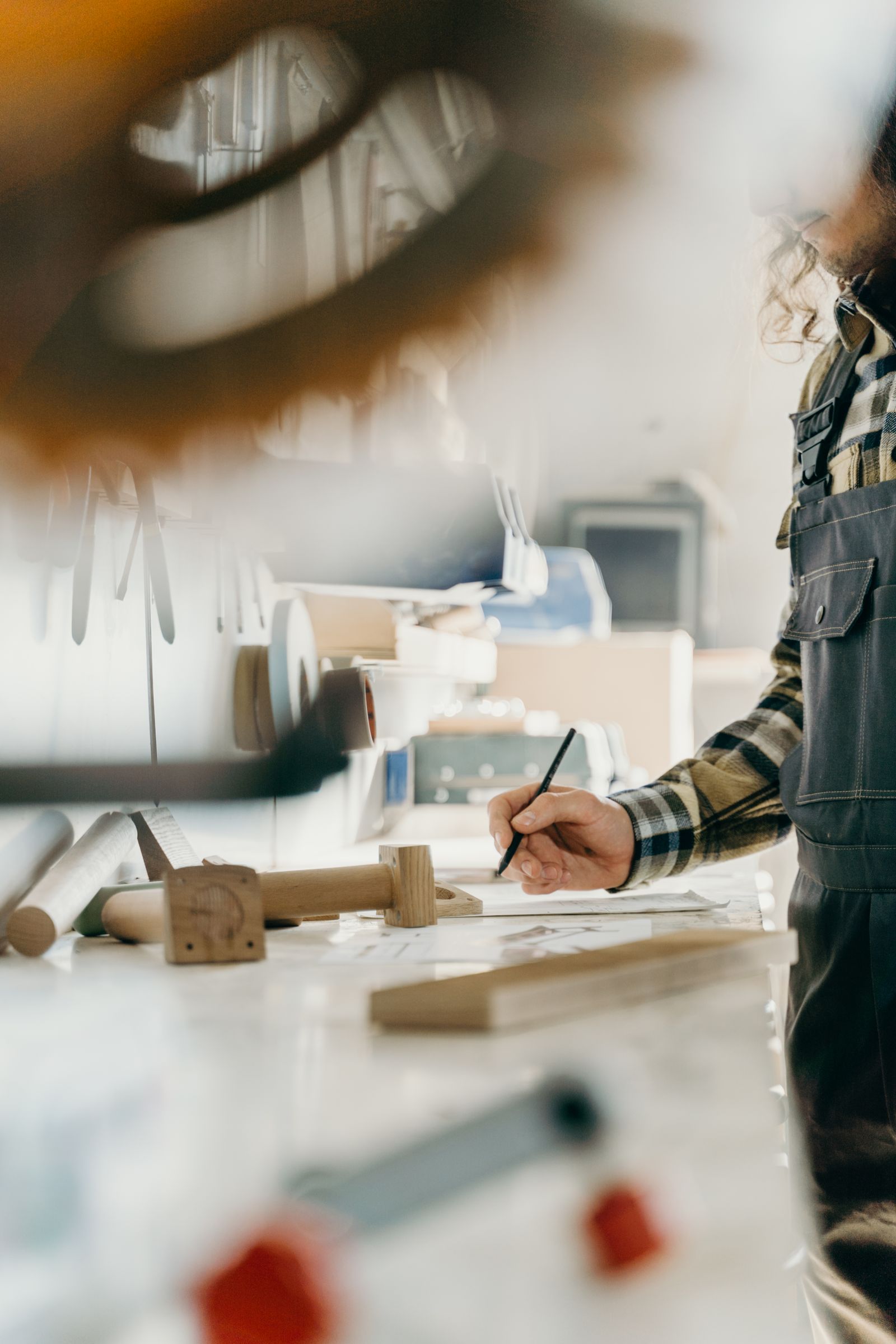 A craftsperson in a bright workshop works at a bench beside woodworking tools, timber components and plans.